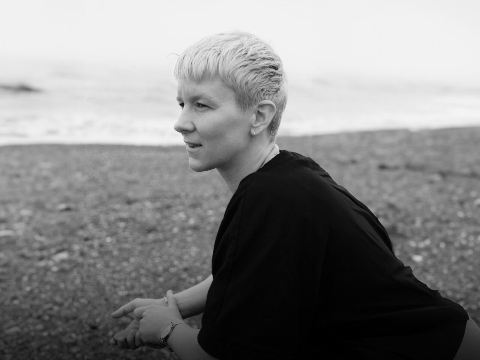 black and white photo of casey, dressed in black, standing on a shingle beach